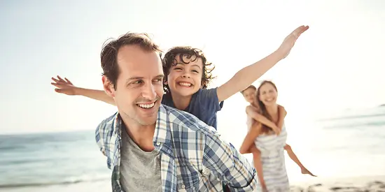 Mutter, Vater und deren zwei Kinder sind am Strand. Die Eltern tragen ihre Kinder Huckepack. 